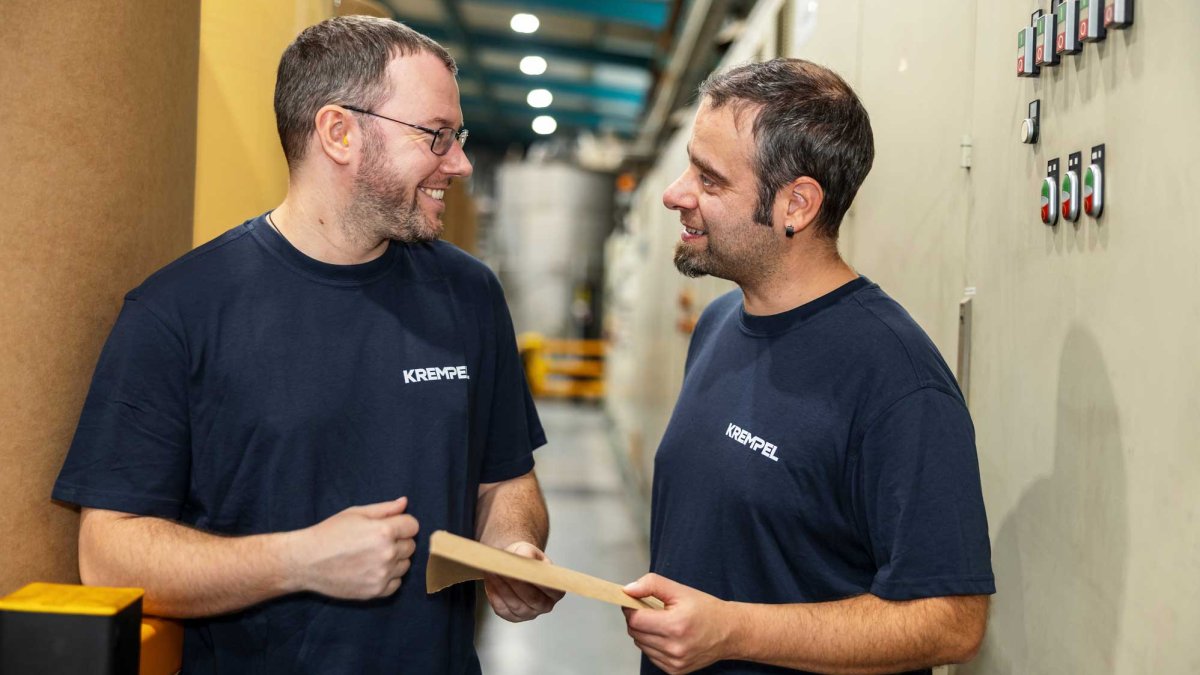 Two men in blue shirts standing in a production hall lookint at each other and talking