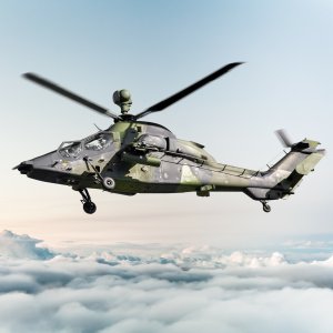 The image shows a military helicopter flying above a layer of clouds.
