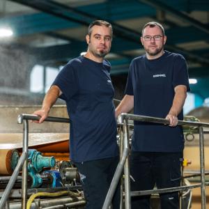 Two men standing in a production area on a platform with steam in the background looking into the camera