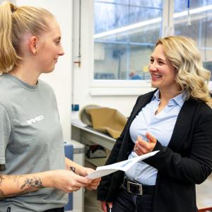 Two young woman standing in a production area looking at each other an discussing a topic