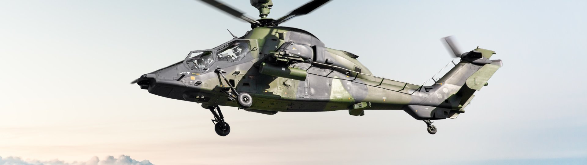 The image shows a military helicopter flying above a layer of clouds.