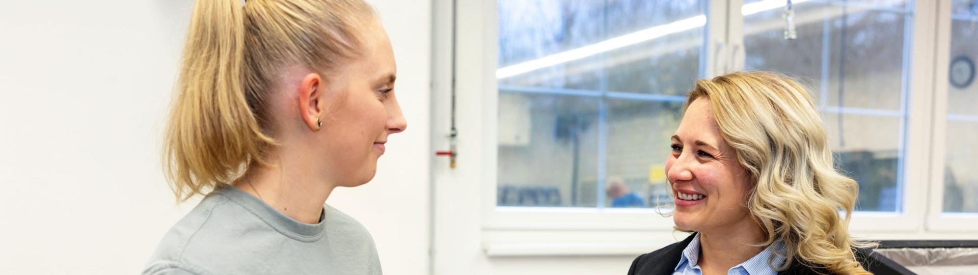 Two young woman standing in a production area looking at each other an discussing a topic