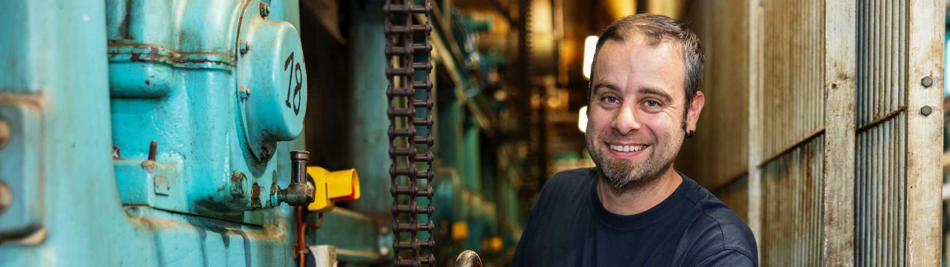 Man working at a machine turning a big wheel and smiling into the camera