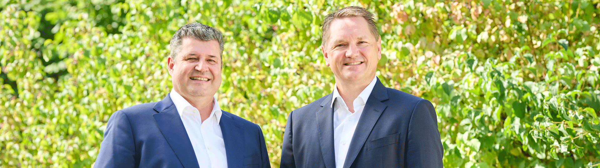 The managing directors of Krempel GmbH are standing in front of a hedge and smiling at the camera.