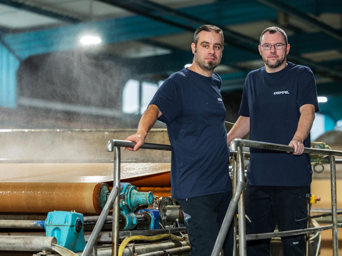Two men standing in a production area on a platform with steam in the background looking into the camera