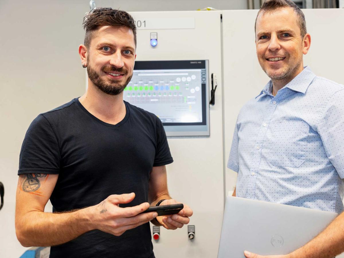 Two men standing next to each other in front of a machine control panel and looking into the camera