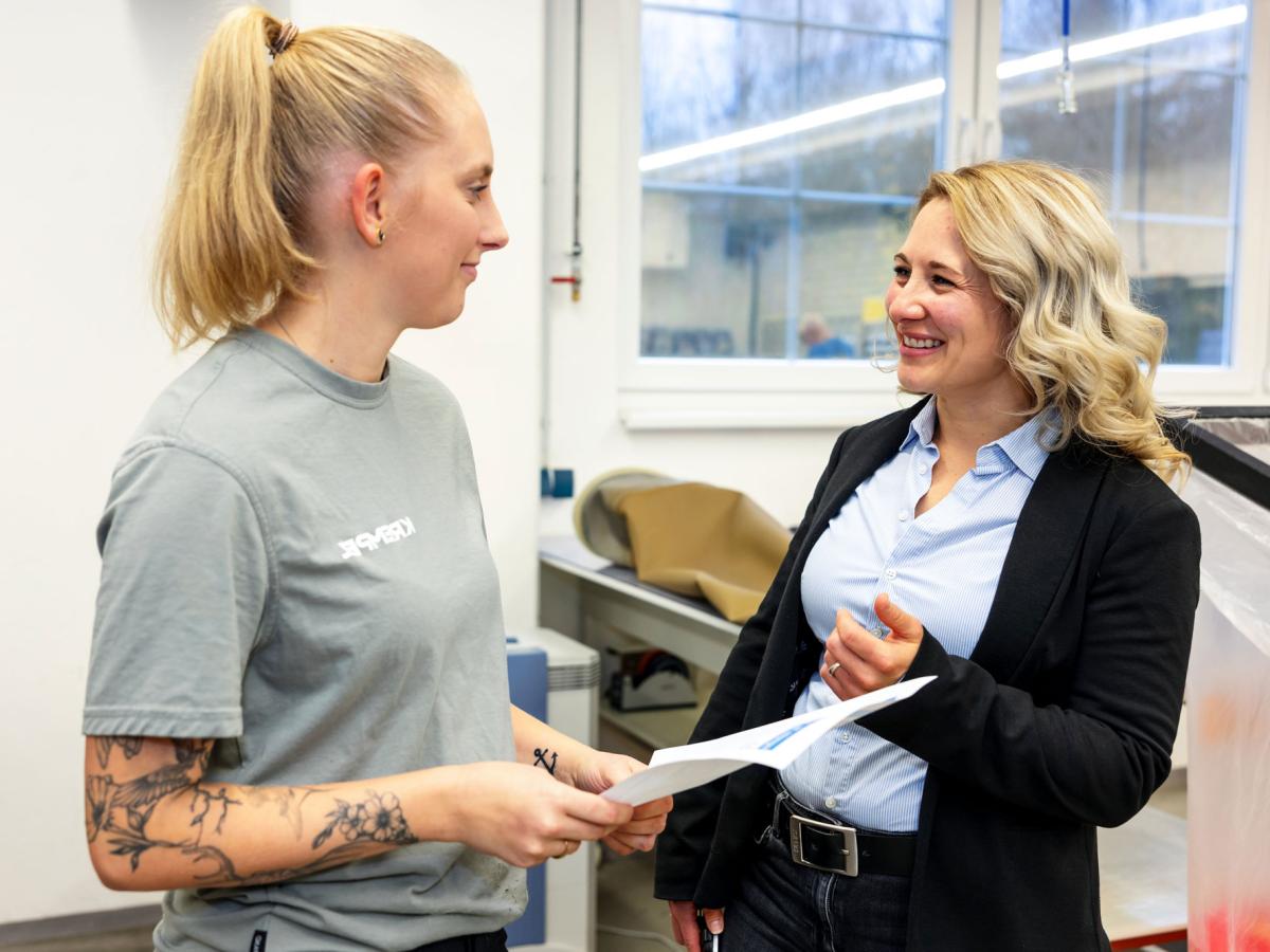 Two young woman standing in a production area looking at each other an discussing a topic