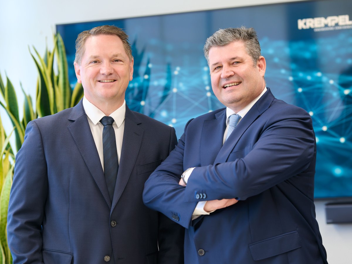The managing directors of Krempel GmbH are standing in front of a screen and a plant, smiling at the camera.