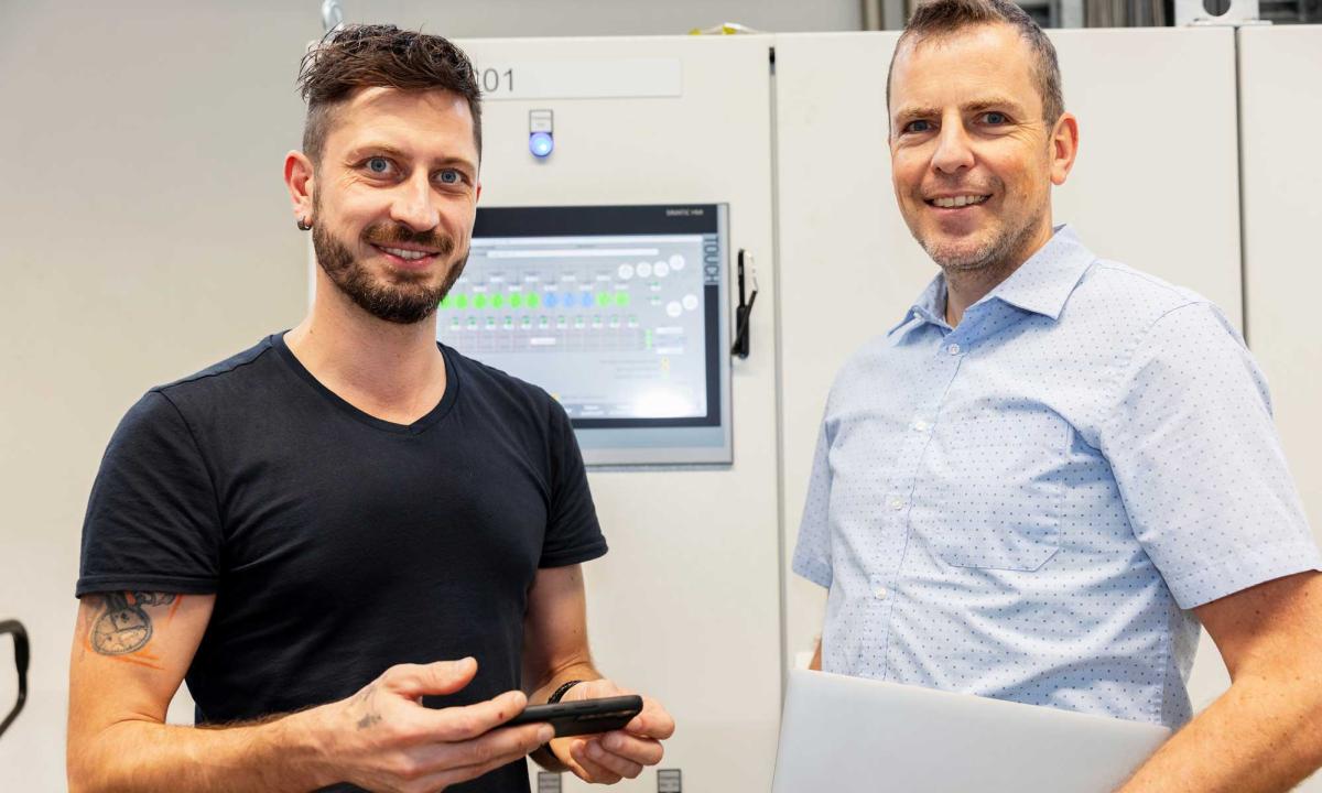 Two men standing next to each other in front of a machine control panel and looking into the camera